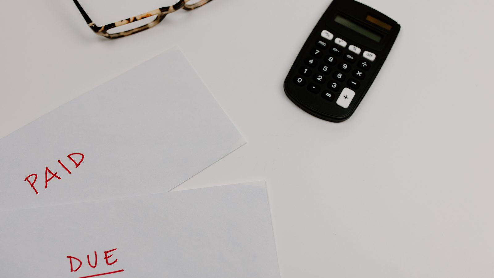 Twee witte enveloppen met de woorden Paid en Due naast een rekenmachine en een bril op een lichte tafel, symbool voor een factuur die nog moet worden betaald